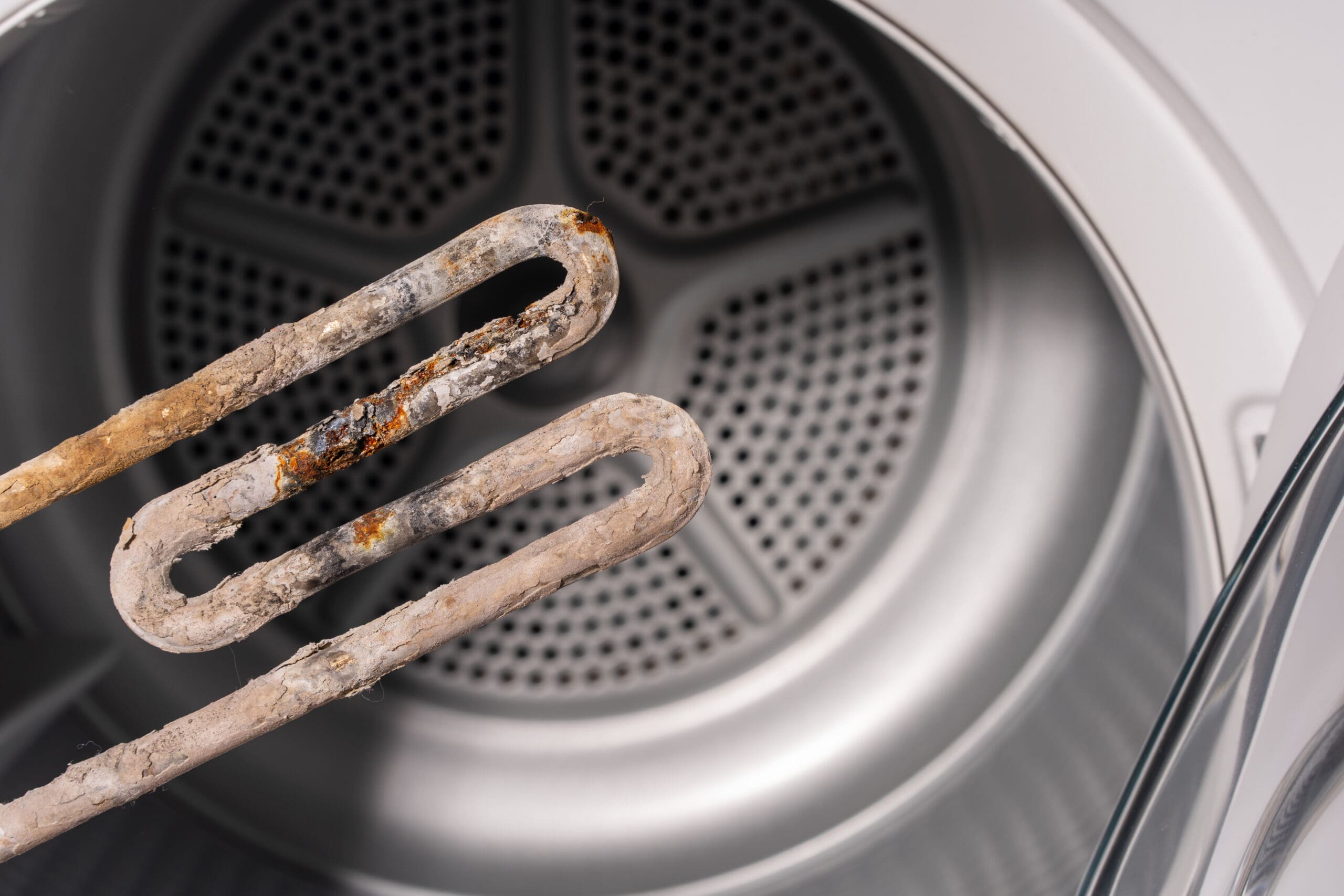 A close-up of a corroded and limescale-covered heating element held in front of an open washing machine drum.
