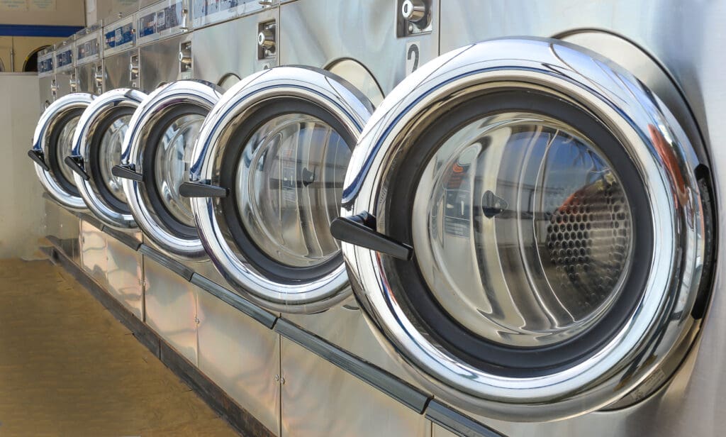 A row of five industrial front-loading washing machines with shiny chrome doors in a laundromat, viewed from an angle. The machines are clean and empty, reflecting light on their metal surfaces.