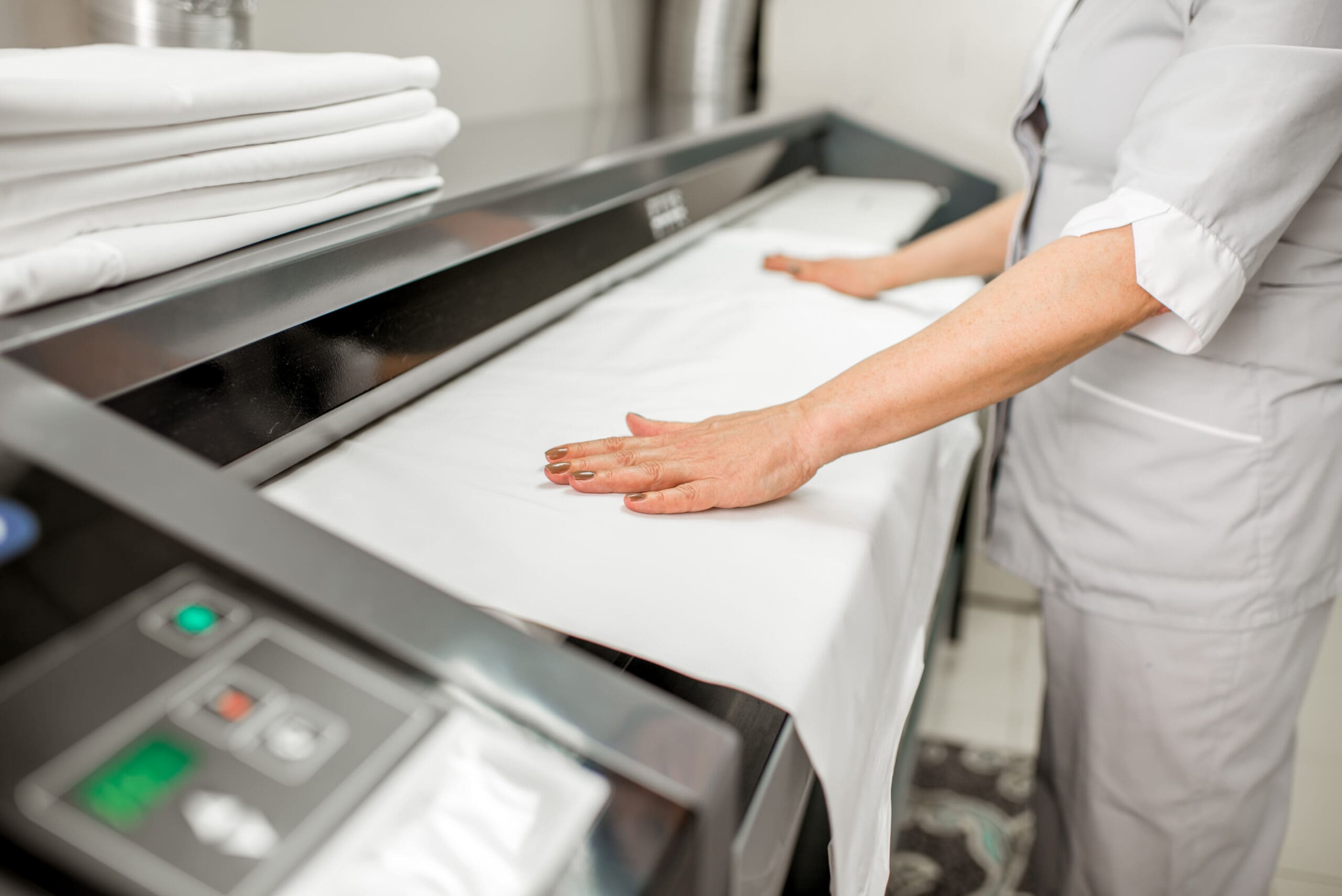 A person in a uniform smooths a white sheet as it passes through a large commercial ironing or pressing machine in a laundry facility, with neatly folded linens stacked nearby.