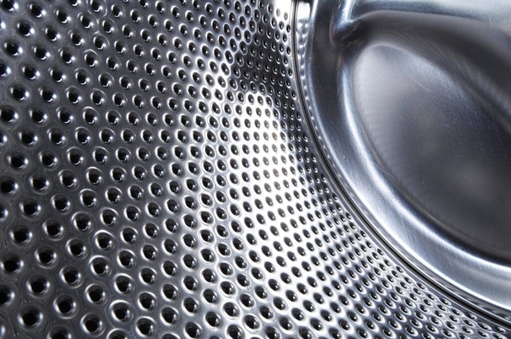 Close-up of the shiny, perforated metal interior drum of a washing machine, showing circular holes and reflective, curved surfaces.