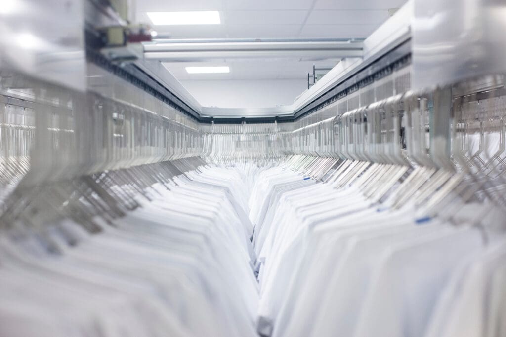Rows of neatly hung white shirts on hangers move along a metal rail system in what appears to be a commercial laundry or dry cleaning facility with bright overhead lighting.