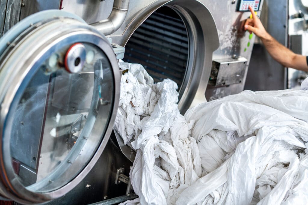 A large industrial washing machine is open, with white linens spilling out, while a person's arm reaches toward the control panel.