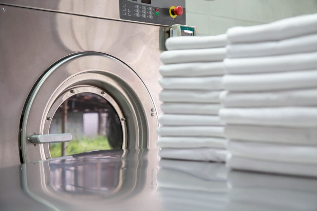 Close-up of a commercial washing machine with a stack of neatly folded white towels on a reflective surface in the foreground. The control panel and part of a tiled wall are visible in the background.