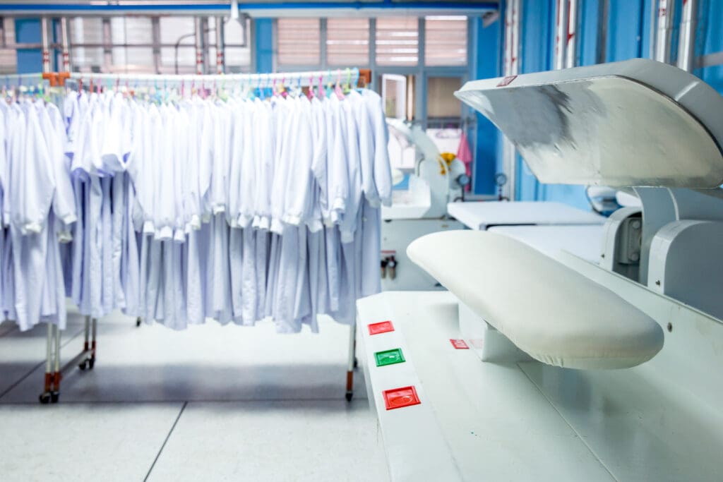 A row of freshly laundered white shirts hangs on racks in a bright, clean laundry facility. In the foreground, there is a large industrial ironing press with red and green buttons.