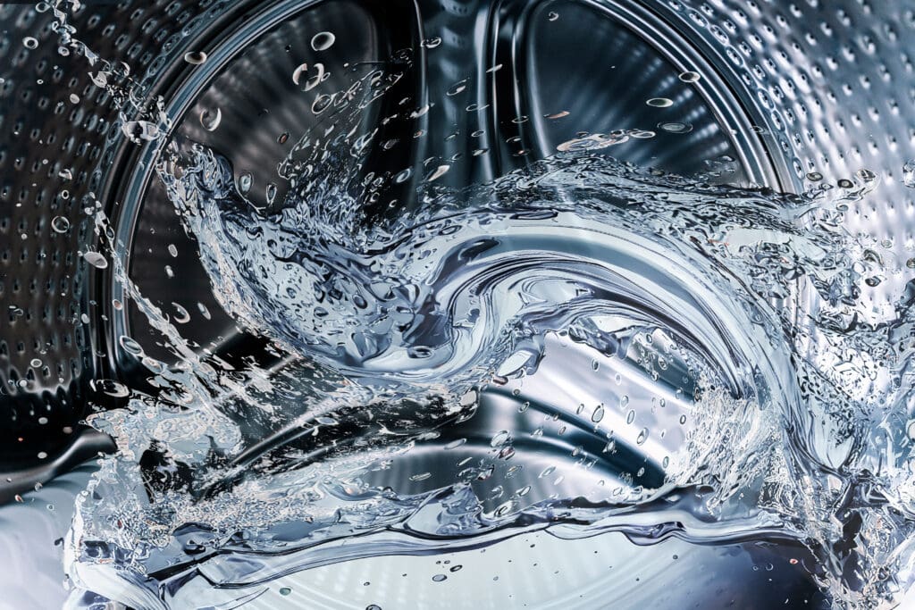 A close-up of swirling water splashing inside a metallic washing machine drum, with droplets and curved reflections creating a dynamic, abstract pattern.