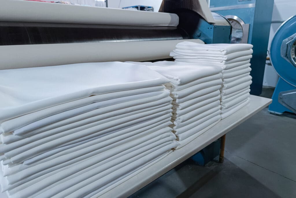 Stacked white linens are neatly arranged on a table in an industrial laundry setting, with large washing and ironing machines visible in the background.