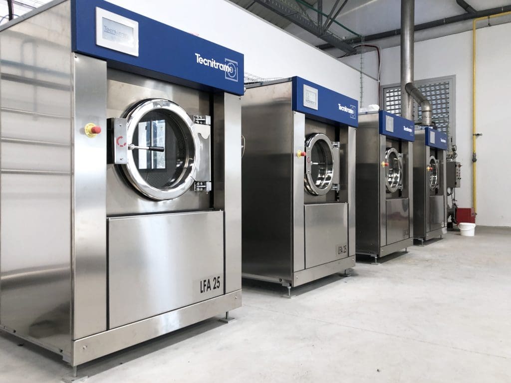 A row of large industrial washing machines with stainless steel exteriors and blue control panels is lined up against a wall in a clean, spacious laundry facility.