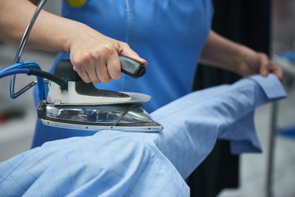 A person in a blue shirt is using a steam iron to press a light blue, checkered dress shirt on an ironing board.