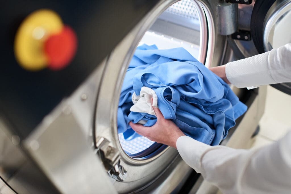 A person places a bundle of blue and white laundry into the open door of an industrial washing machine.