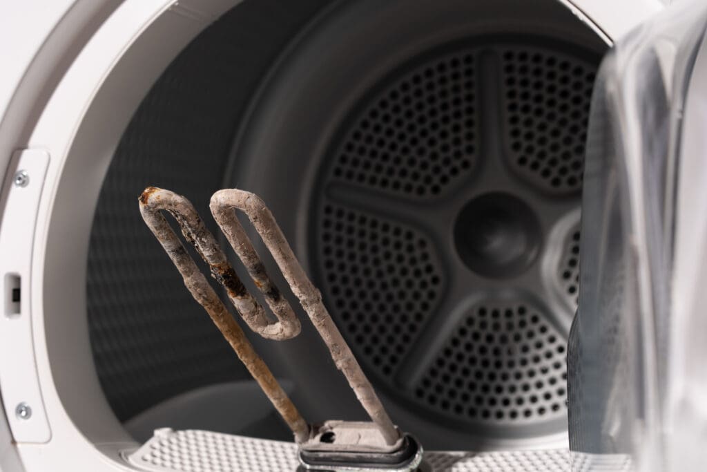 A rusty heating element is held in front of an open clothes dryer drum, highlighting damage or wear to the appliance part.