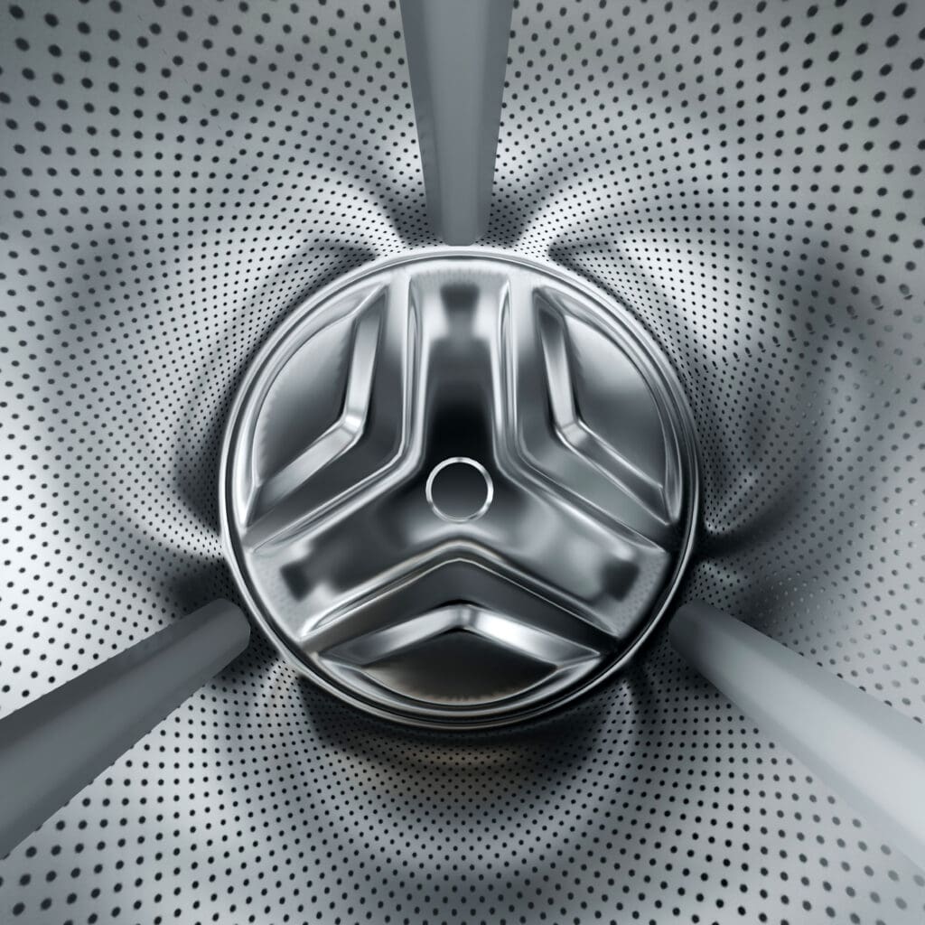 Close-up view of the inside drum of a washing machine, showing a circular metal pattern with many small holes and three prominent metal fins extending from the center.