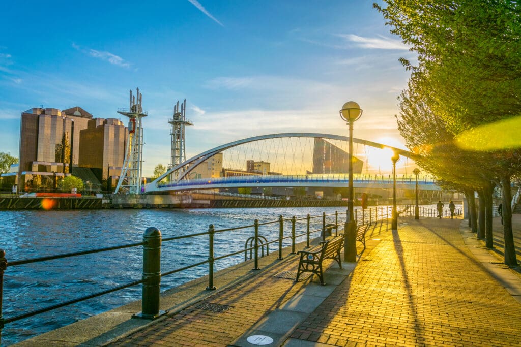 A riverside walkway at sunset with modern buildings, a curved bridge, and trees lining the path. Sunlight creates a warm glow and long shadows on the pavement.