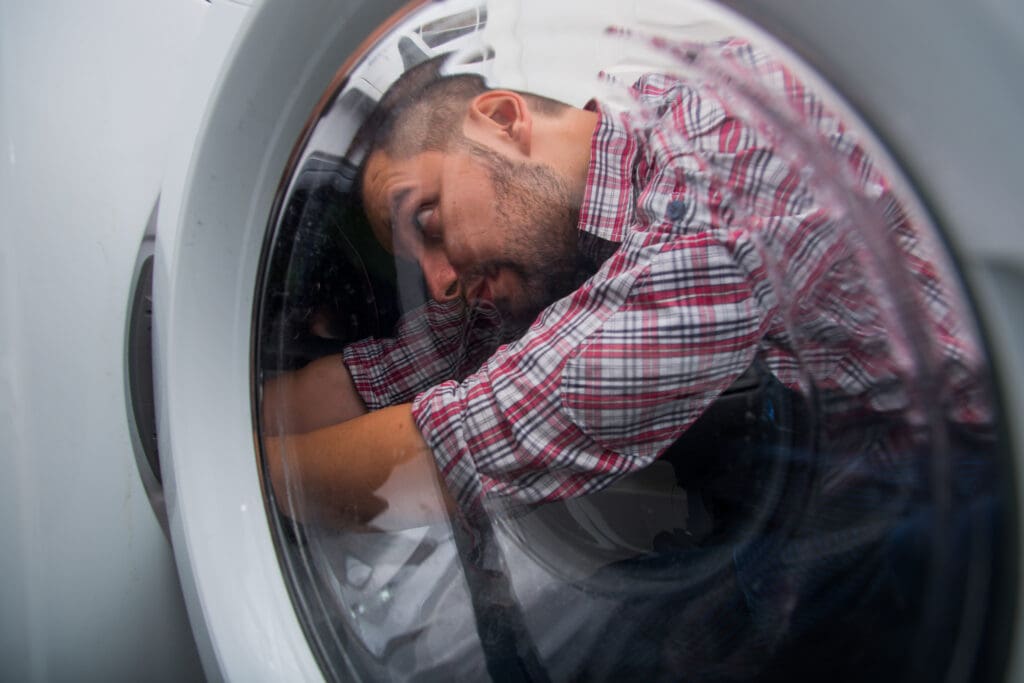 A man in a plaid shirt is curled up inside a washing machine, viewed through the glass door, with his head resting on his arm and his eyes closed.