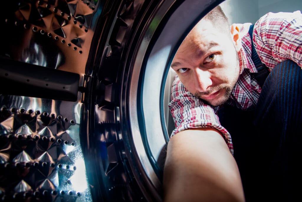 A man in a plaid shirt and suspenders is reaching into the open door of a washing machine, photographed from inside the drum, with light illuminating his face and the metallic interior.