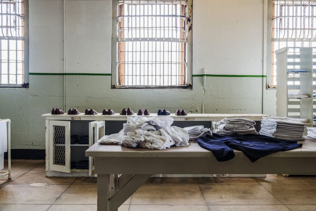 A room with barred windows, stacks of folded clothes, and shoes lined up on shelves, resembling a prison laundry or uniform distribution area.