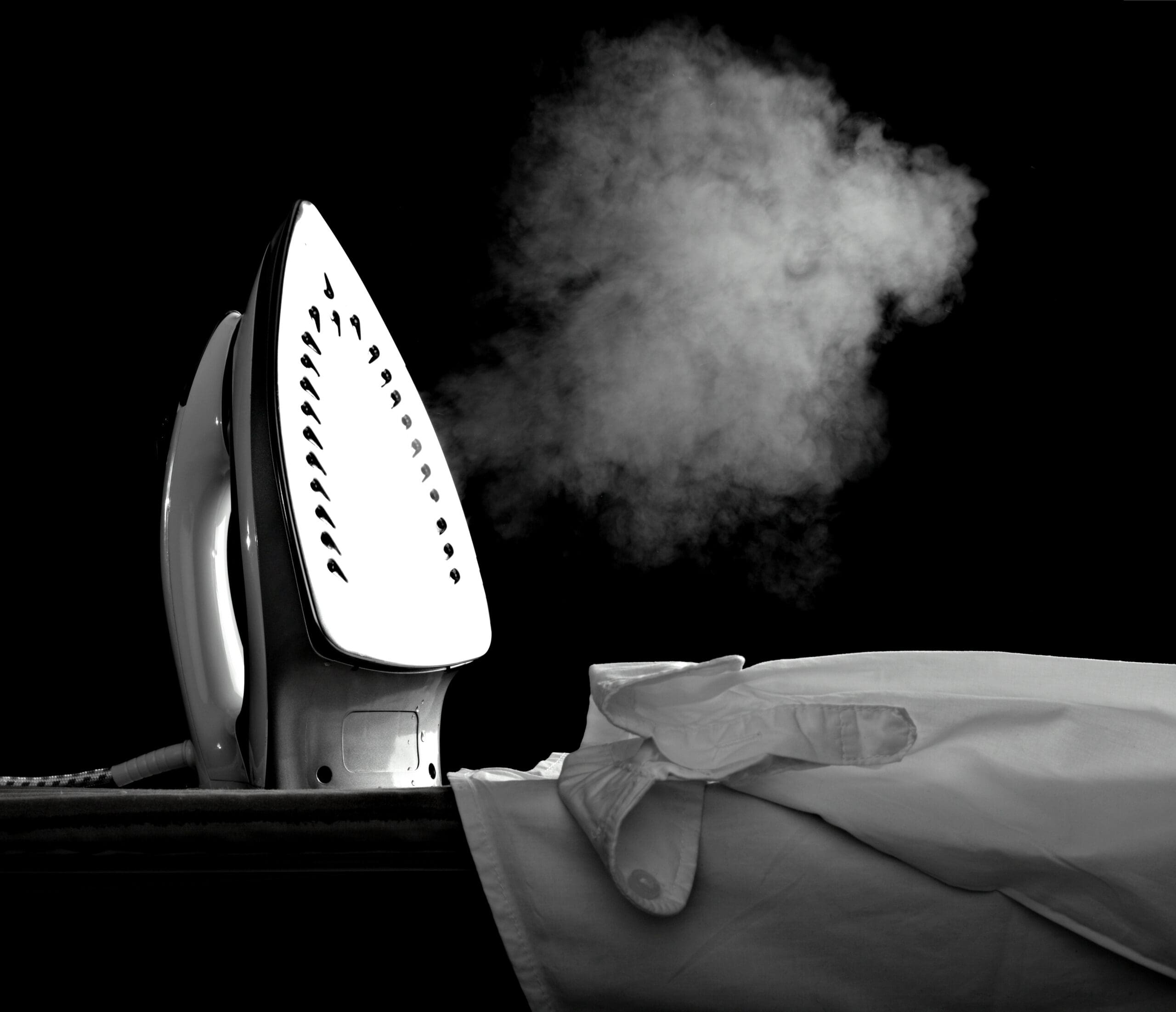 A steam iron releases a cloud of steam above a white dress shirt on an ironing board, set against a black background.