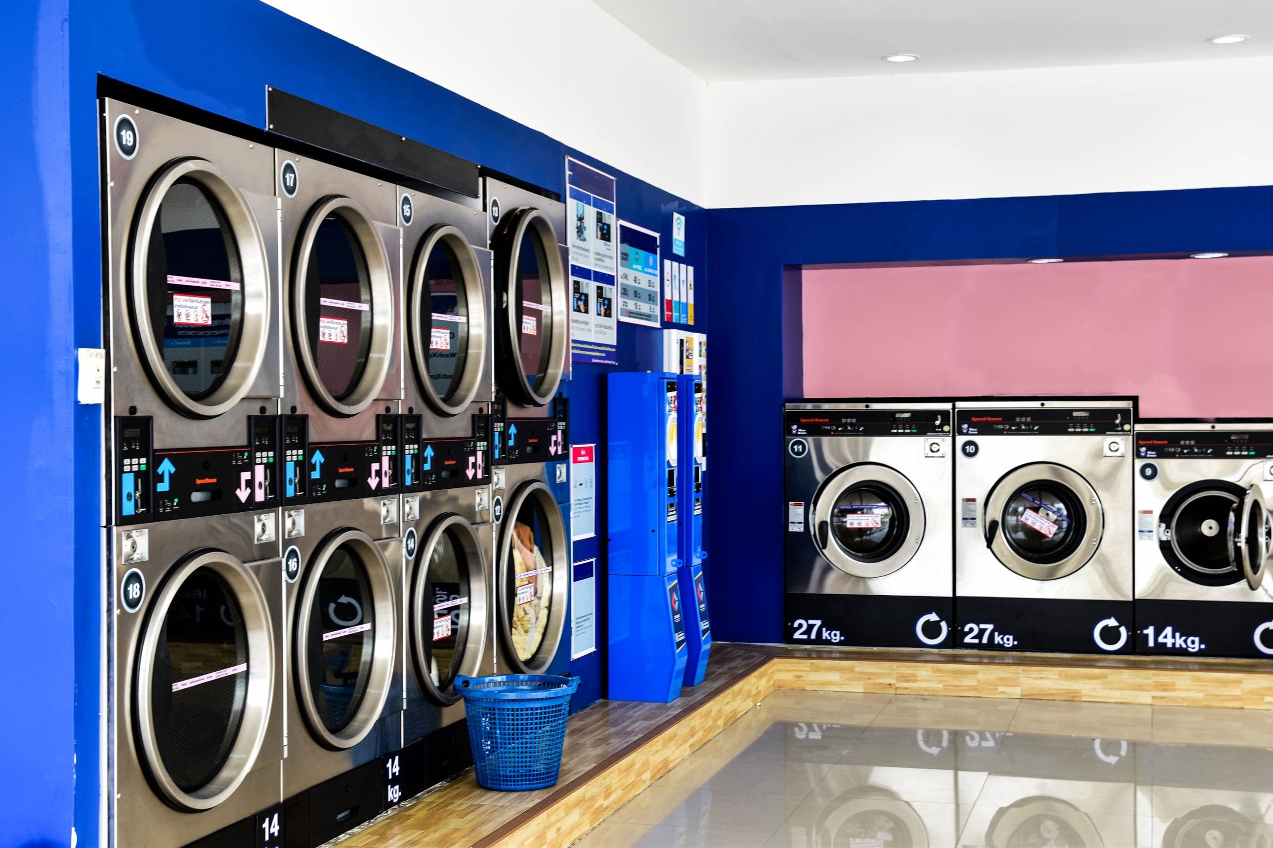 Rows of large front-loading washing machines and dryers in a brightly lit self-service laundromat, with blue walls, signage, and a blue laundry basket on the floor.