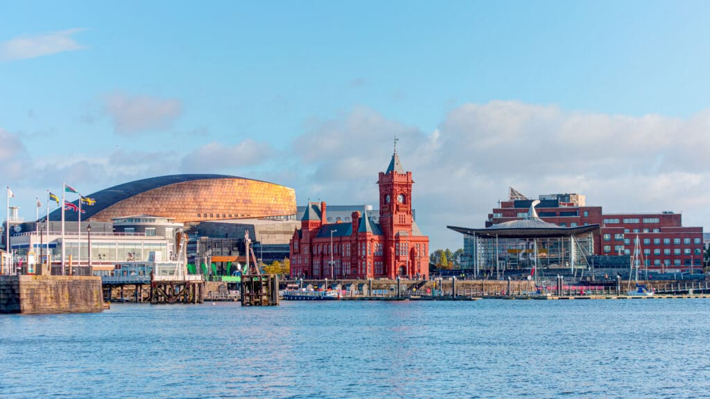 Waterfront view of Cardiff Bay with the red-brick Pierhead Building, the Wales Millennium Centre with its copper dome, and modern buildings under a blue sky with scattered clouds.