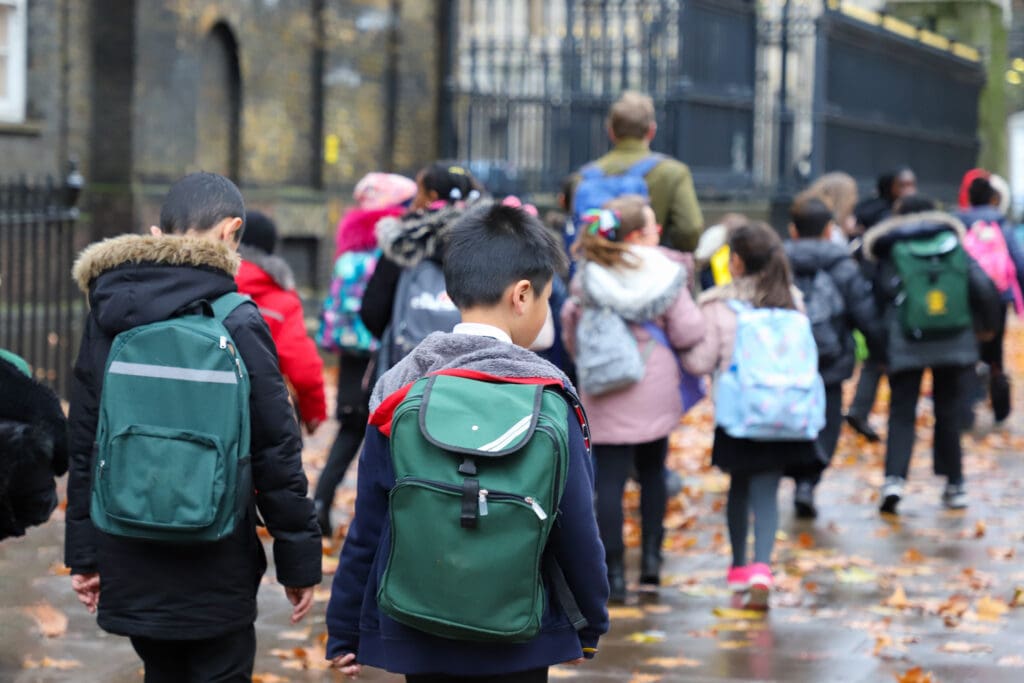 A group of schoolchildren wearing backpacks walk along a wet, leaf-covered sidewalk, accompanied by an adult, near a black iron fence and old brick building.