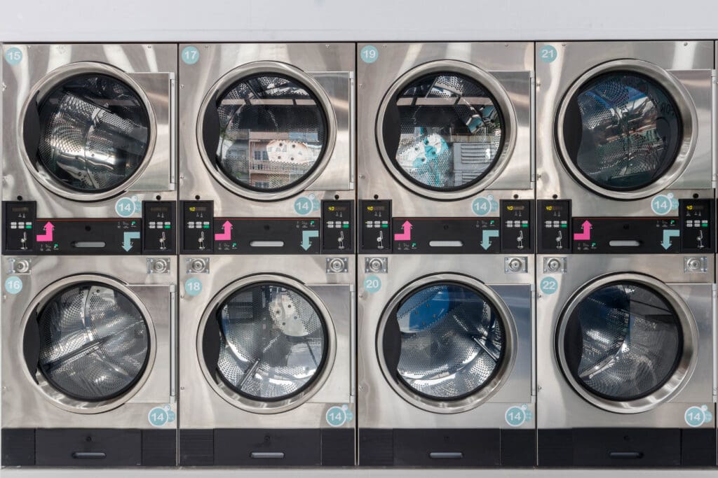 Eight industrial washing machines and dryers arranged in two rows, with shiny stainless steel doors and digital control panels, are seen in a clean, modern laundromat setting.