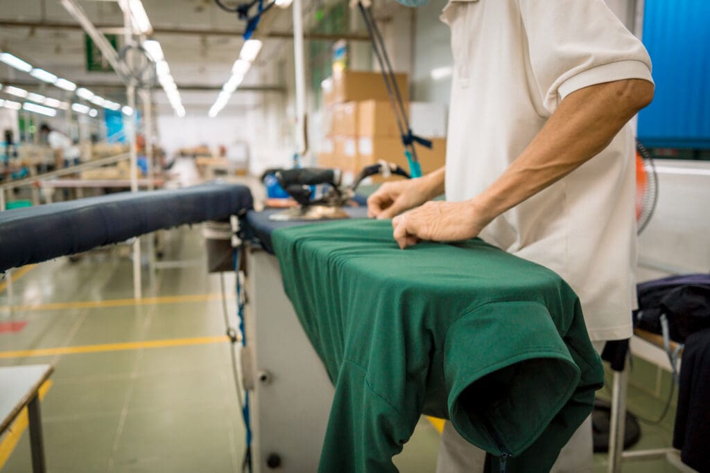 A person in a white shirt irons a green garment on an ironing board in a brightly lit factory or workshop. The background shows machinery, boxes, and a spacious work area.
