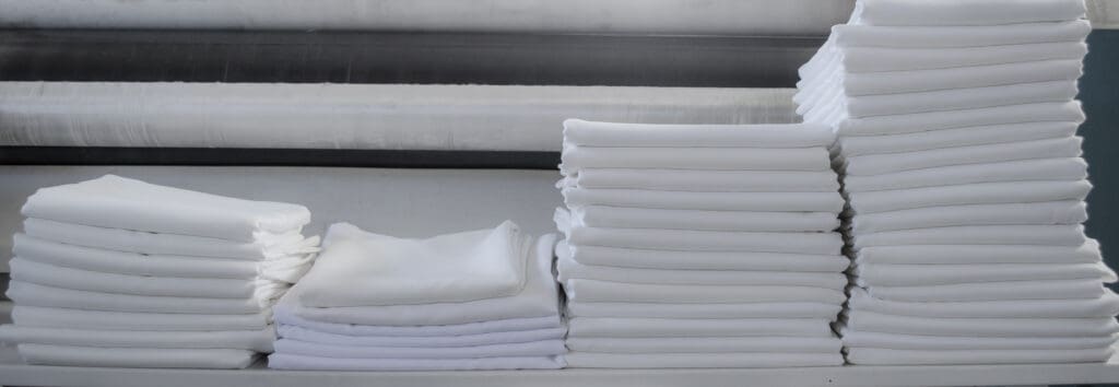 Stacks of neatly folded white linens are arranged in varying heights on a flat surface, with a fabric roll visible in the background.
