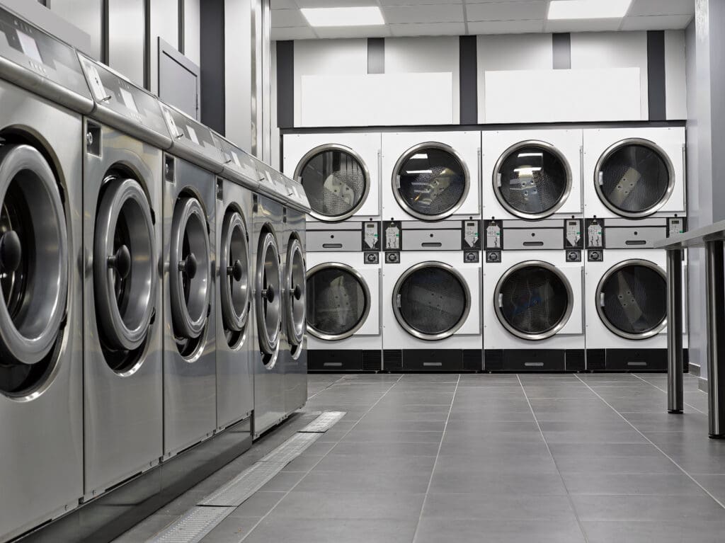 A row of front-loading washing machines and dryers in a modern, empty laundromat with gray tiled floors and white walls.