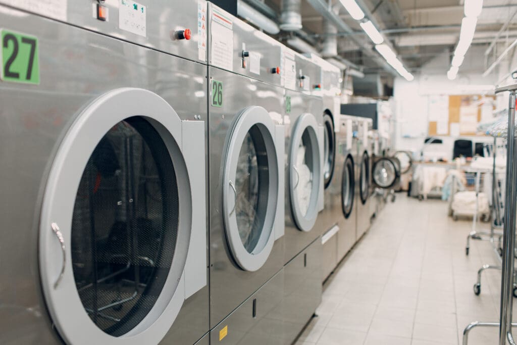 A row of large industrial washing machines in a brightly lit laundromat. The floor is tiled, and laundry carts are visible in the background. The machines are silver with numbered labels.