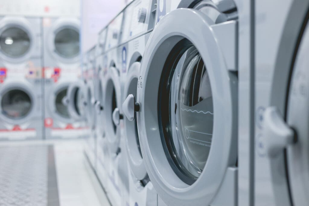 A close-up view of a row of modern front-loading washing machines in a laundromat, with a focus on one machine’s door and others lined up in the background.