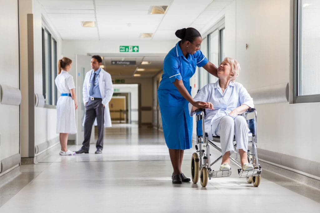 A nurse smiles and talks to an elderly woman sitting in a wheelchair in a hospital corridor. In the background, two medical staff stand together and talk. The hallway is bright and clean with large windows.