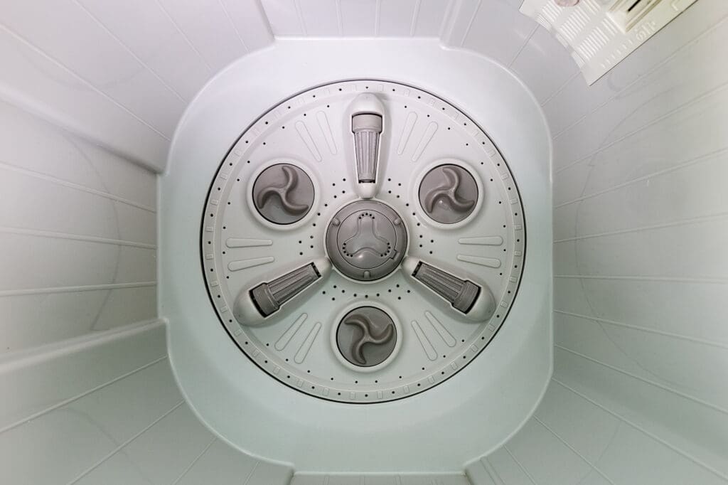 The image shows the inside of a top-loading washing machine drum, viewed from above, with a central agitator and evenly spaced fins around it. The drum is clean, white, and empty.