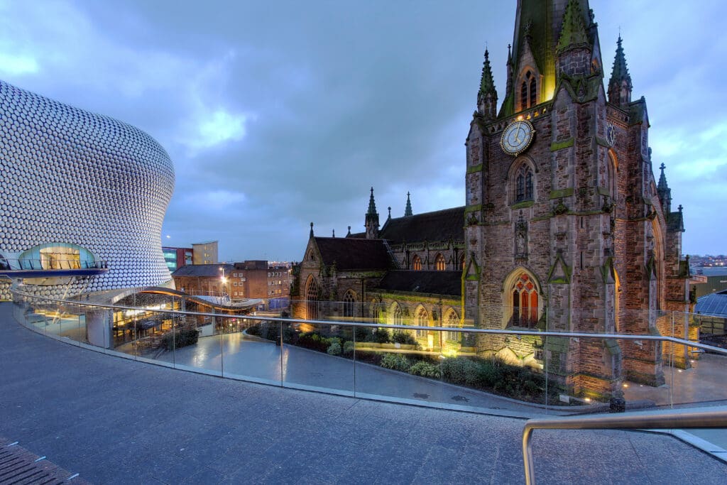 A view of St Martin's Church in Birmingham, UK, with its illuminated clock tower, next to the modern, curved facade of the Selfridges building at dusk under a cloudy sky.