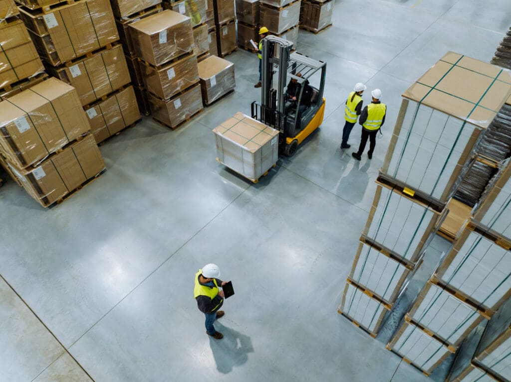 Aerial view of warehouse workers in safety vests and helmets. A forklift moves a pallet while two workers stand nearby and another worker checks a clipboard among large stacks of boxes and pallets.