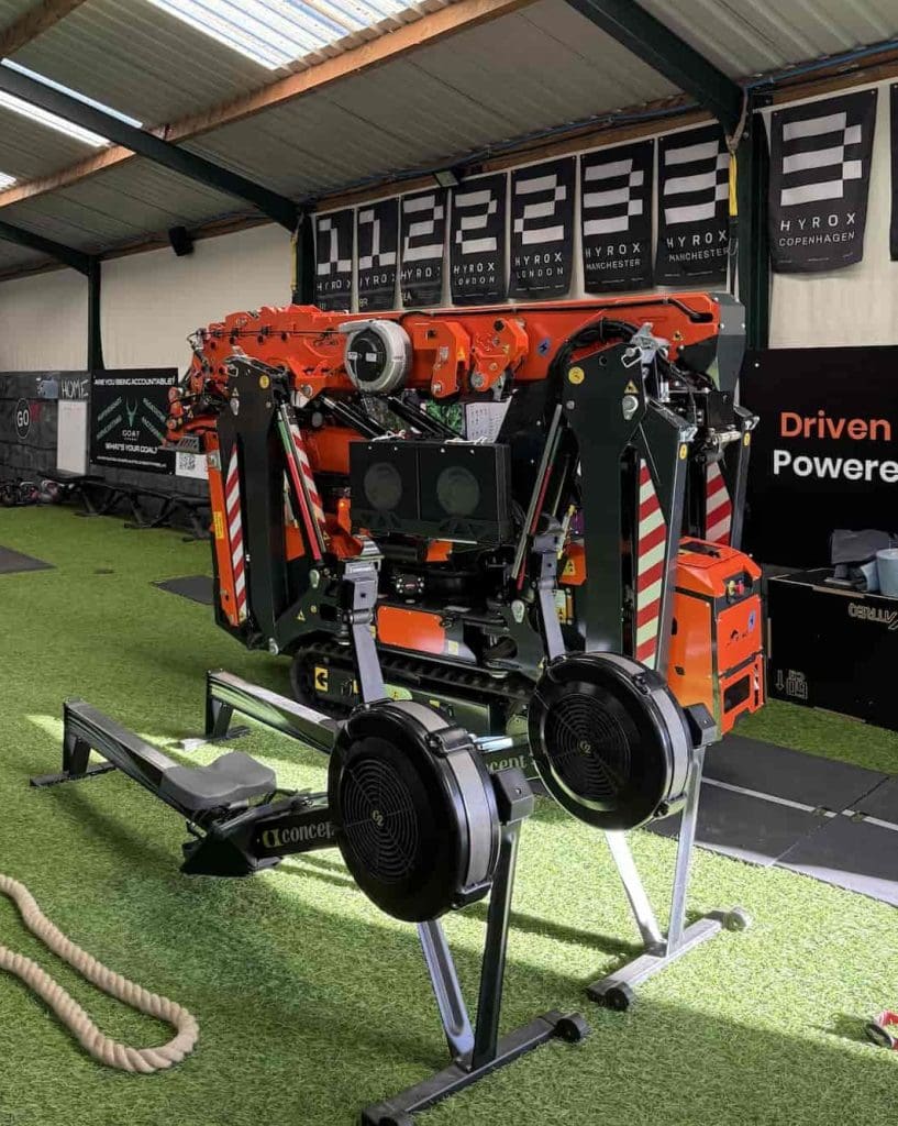 A large, complex orange and black machine is set up indoors on artificial turf, surrounded by gym equipment like a rowing machine and ropes, under a corrugated metal roof with black HYROX banners on the wall behind.