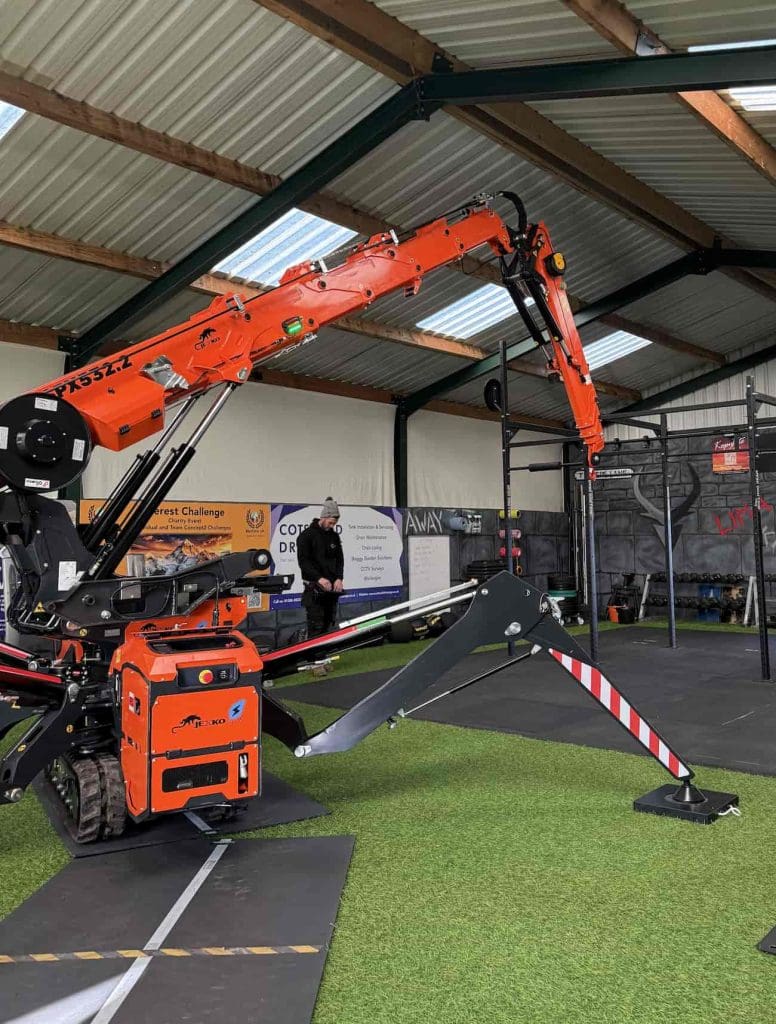 A compact orange spider crane with extended stabilizer legs is positioned on artificial turf inside a building. A person in dark clothing stands nearby, and gym equipment and banners are visible in the background.